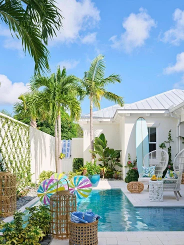 Bright modern white pool house with palm trees, a surfboard, and woven outdoor furniture by a turquoise swimming pool.